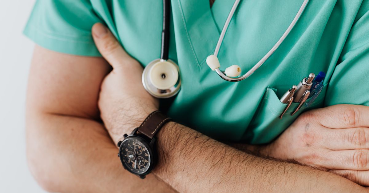 Unrecognizable man wearing medical uniform with stethoscope and pens standing in hospital with crossed arms during work in day time